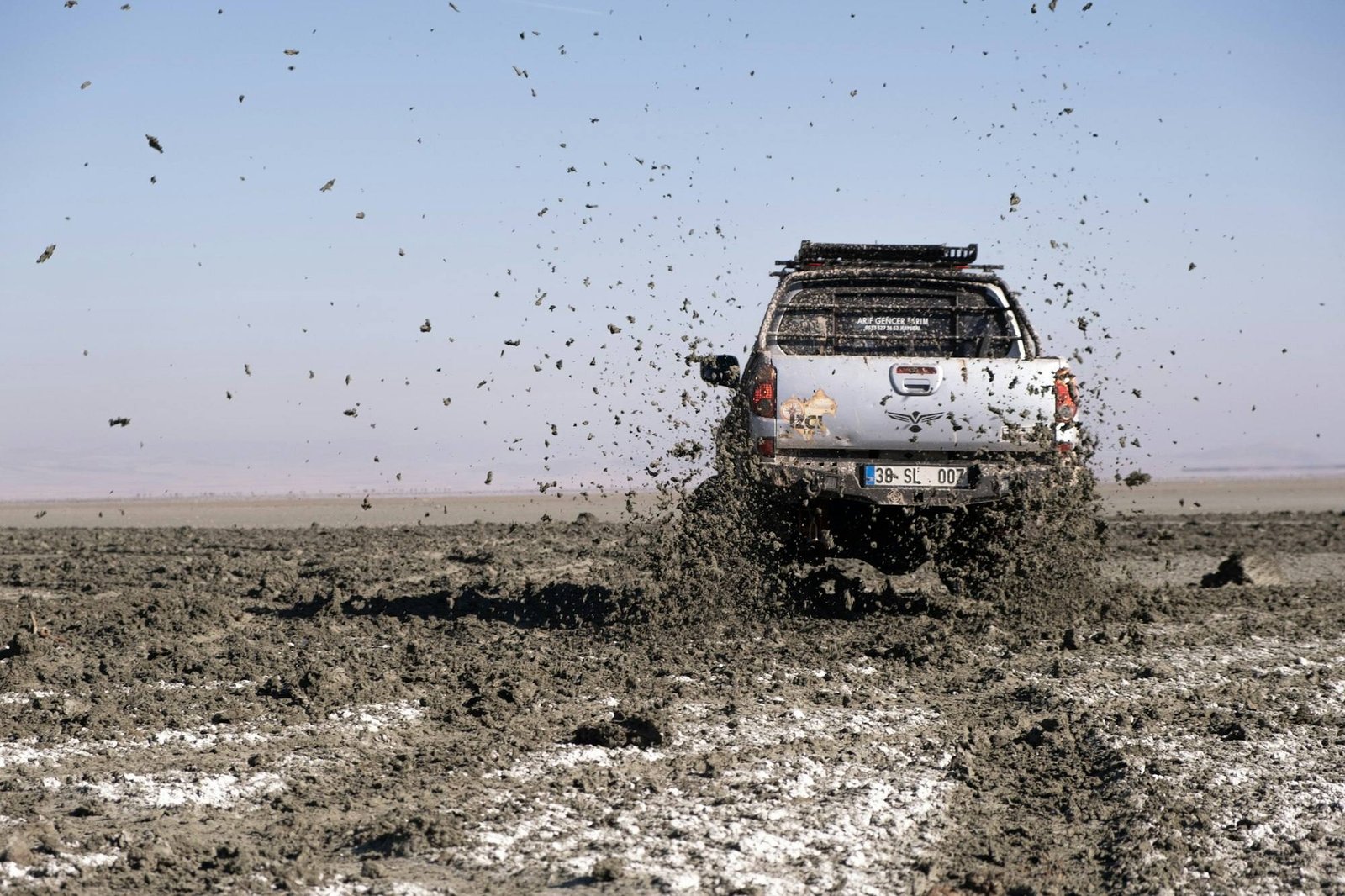 A pickup with a roof rack driving through deep mud on an open flat, mud flying behind it
