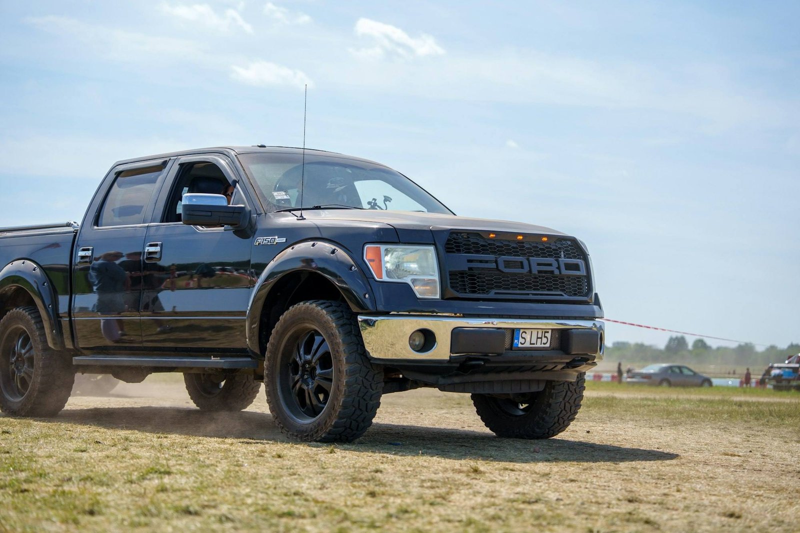 A dark Ford F-150 pickup on off-road wheels driving across a dusty field