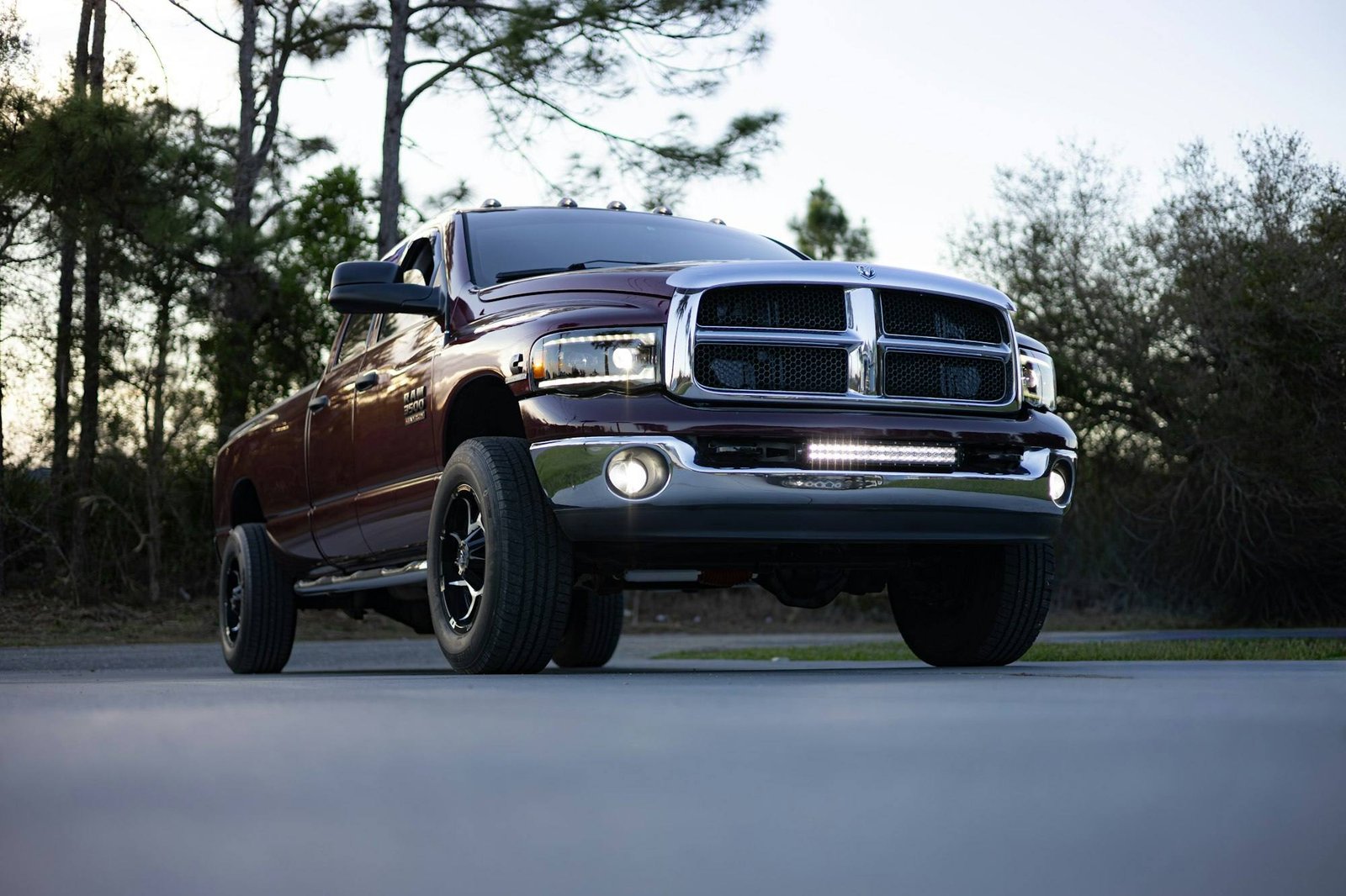 A lifted maroon Ram pickup on large off-road wheels parked beside a road lined with pine trees