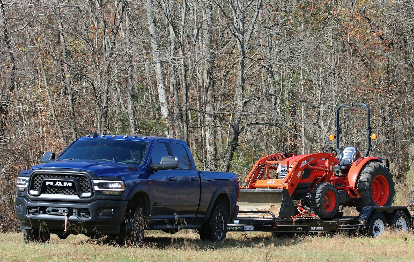 A blue Ram 2500 Power Wagon hauling a trailer with a compact tractor past bare winter trees