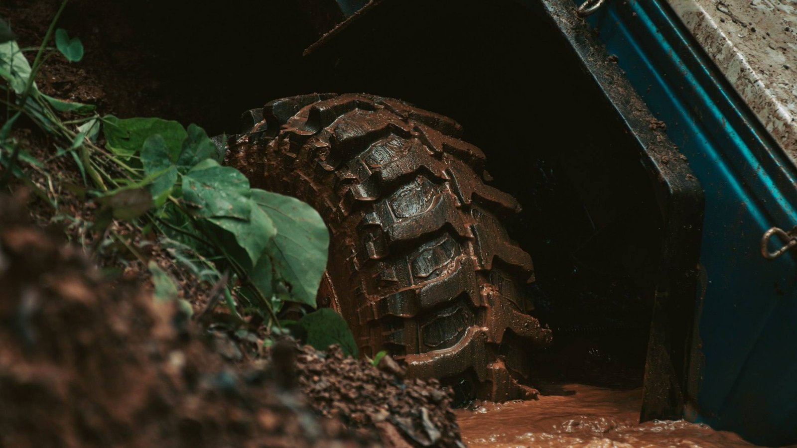 A deep-lugged off-road tire caked in dark mud as it splashes through brown water