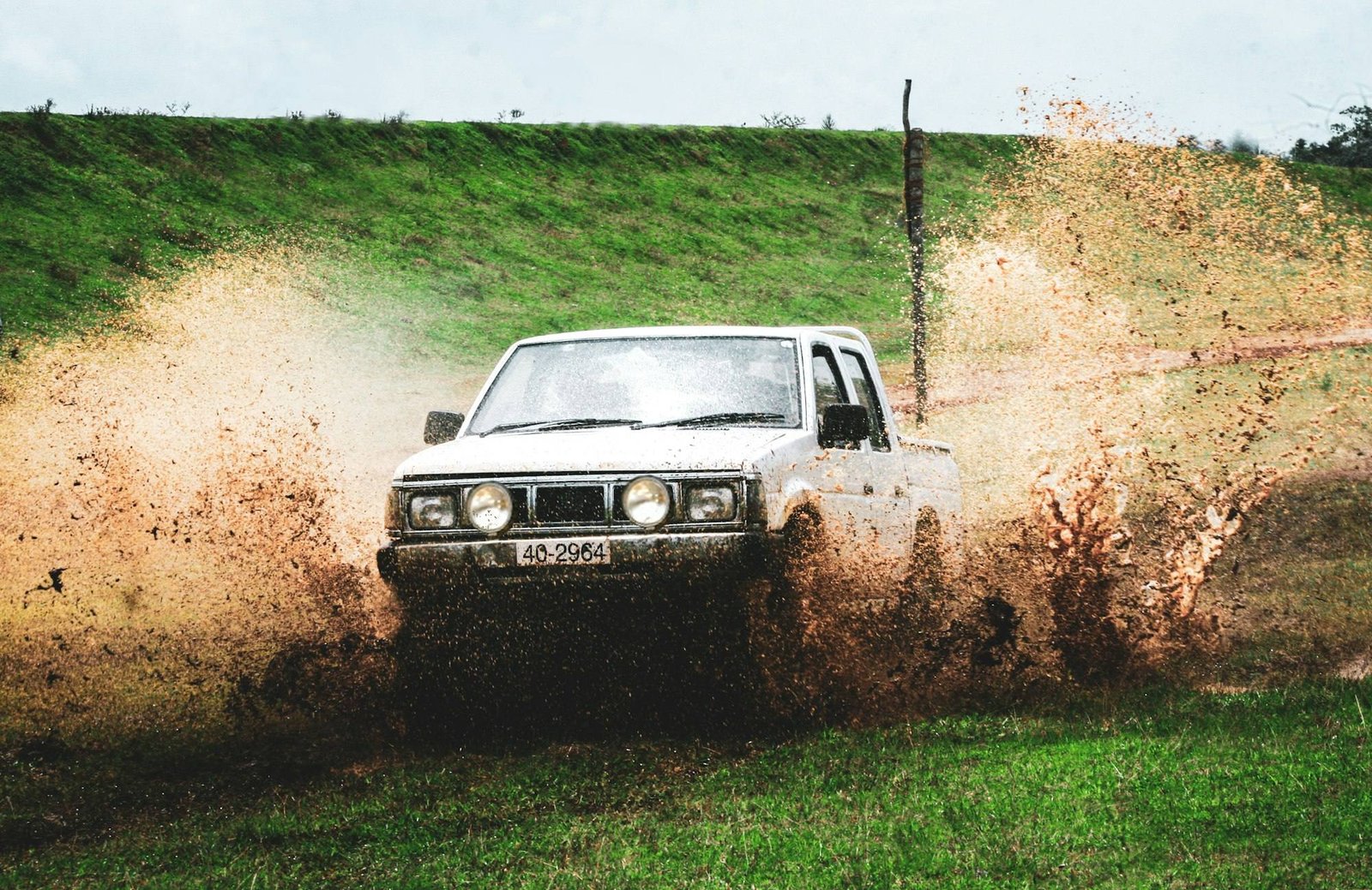A white pickup splashing through a muddy puddle beside a grassy bank